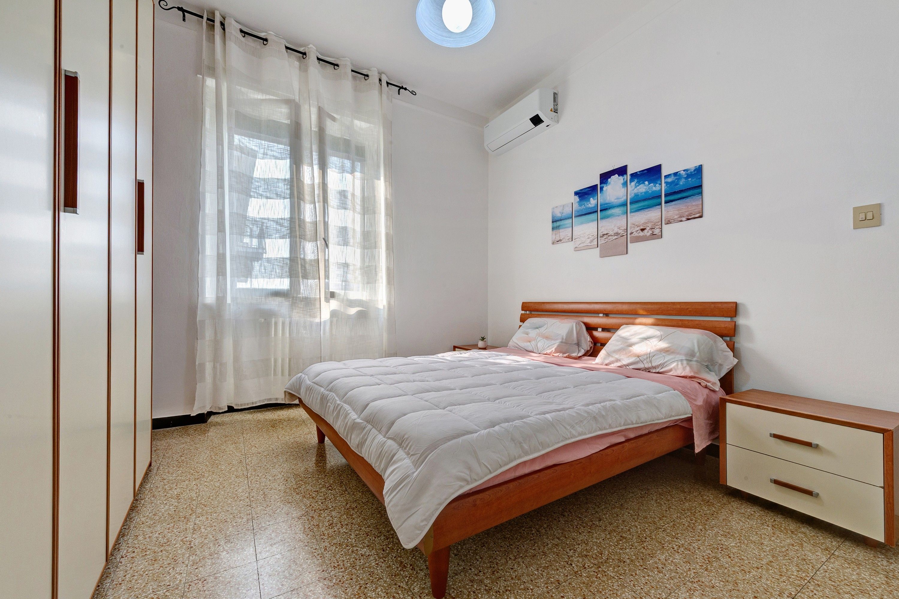 A bedroom with a bed, a nightstand, a wall-mounted air conditioner, and a window with sheer curtains.