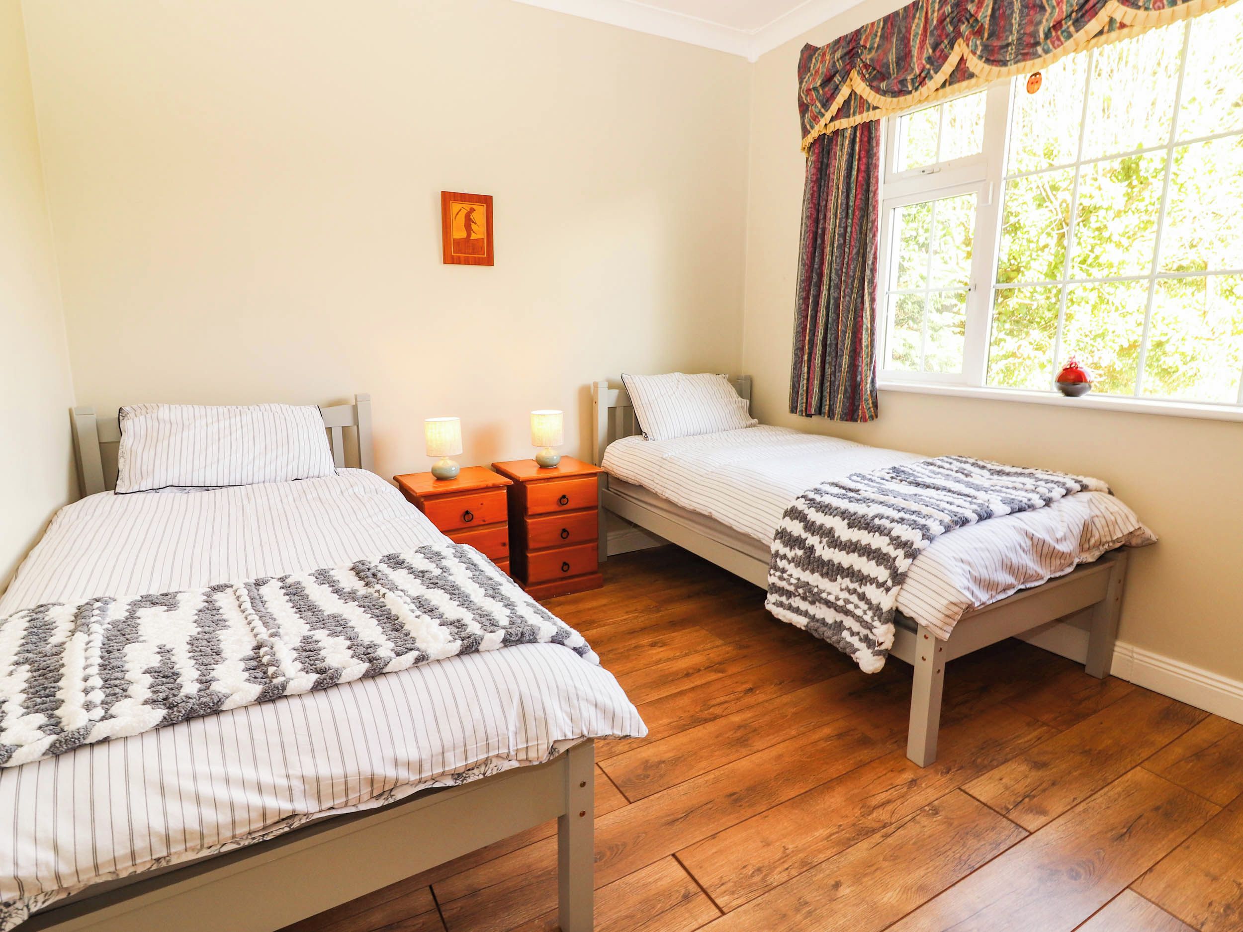 A bedroom with two single beds, wooden nightstands with lamps, a window with curtains, and a wall-mounted picture.