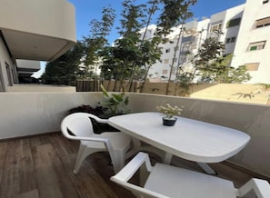Een balkon met een witte tafel en stoelen, een potplant en uitzicht op omliggende gebouwen en bomen.