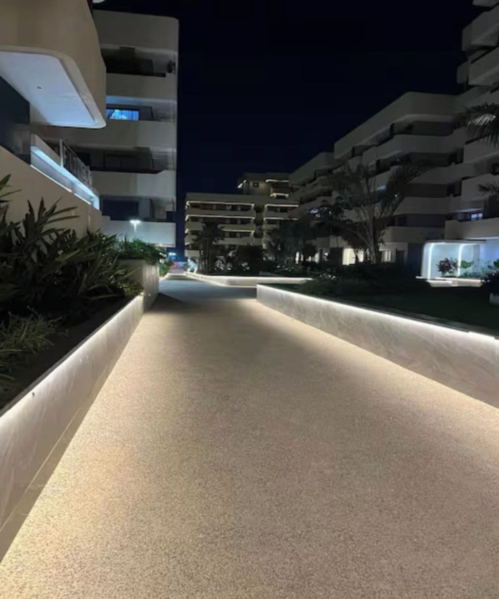 A nighttime view of a modern urban street with illuminated pathways, multi-story buildings, and landscaped areas.
