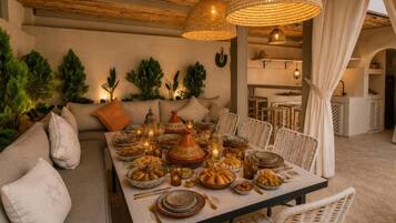 A dining area with a long table set for a meal, surrounded by wicker chairs, and a hanging light fixture above.