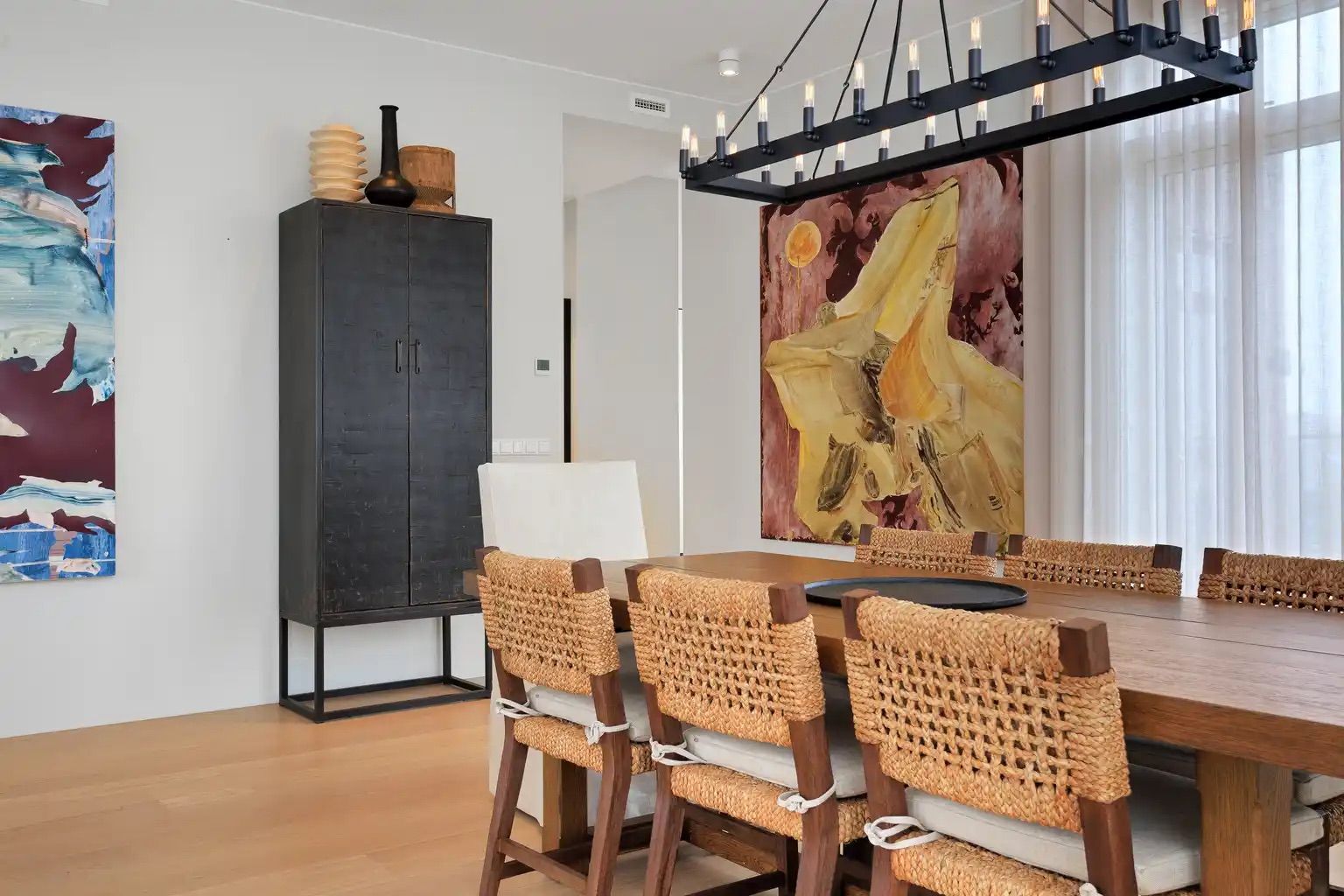 A modern dining area with a wooden table, woven chairs, a dark cabinet, and a large abstract painting.
