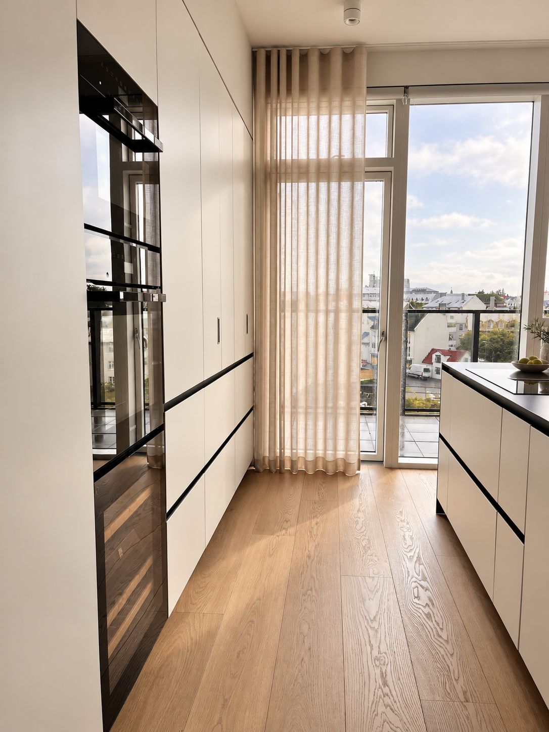 A modern kitchen with wooden flooring, white cabinets, and a view of the city through a glass door.