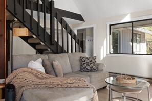 A modern living room with a grey sofa, a woven throw blanket, a glass coffee table with a woven basket, and a black staircase. - Lakeview Retreat + Boat Slip & Waterfront Access (Coeur d'Alene)