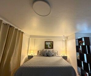 Un dormitorio con cama, dos mesitas de noche, un cuadro en la pared y una estantería con flores.