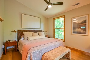 A bedroom with a bed, a bedside table, a nightstand, a ceiling fan, a window with a view of trees, and a framed picture on the wall. - Ramblewood Lodging 5 (Athens)
