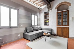 A modern living room with a grey sectional sofa, a small round table, and a white rug. There are two windows, a wooden cabinet with glass doors, and a shelf unit against the wall. - Bombarde Stone and Beams (Florence)