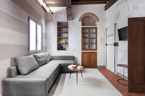 A modern living room with a grey sectional sofa, a small round table, and a built-in shelving unit with glass doors. - Bombarde Stone and Beams (Florence)