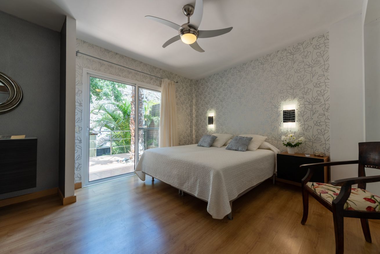 Een slaapkamer met een bed, een plafondventilator, een nachtkastje, een stoel en een raam met uitzicht op groen.