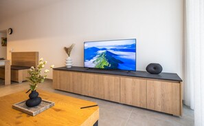 A modern living room with a wooden cabinet, a flat-screen TV displaying a mountain landscape, a vase with flowers, and a wooden coffee table. - Charming 1BR Getaway, Great for Couples (Msida)