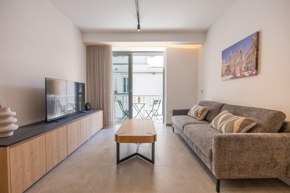 Ein modernes Wohnzimmer mit Sofa, Couchtisch, Fernseher auf einer Holzkonsole und einem Balkon mit Tisch und Stühlen.