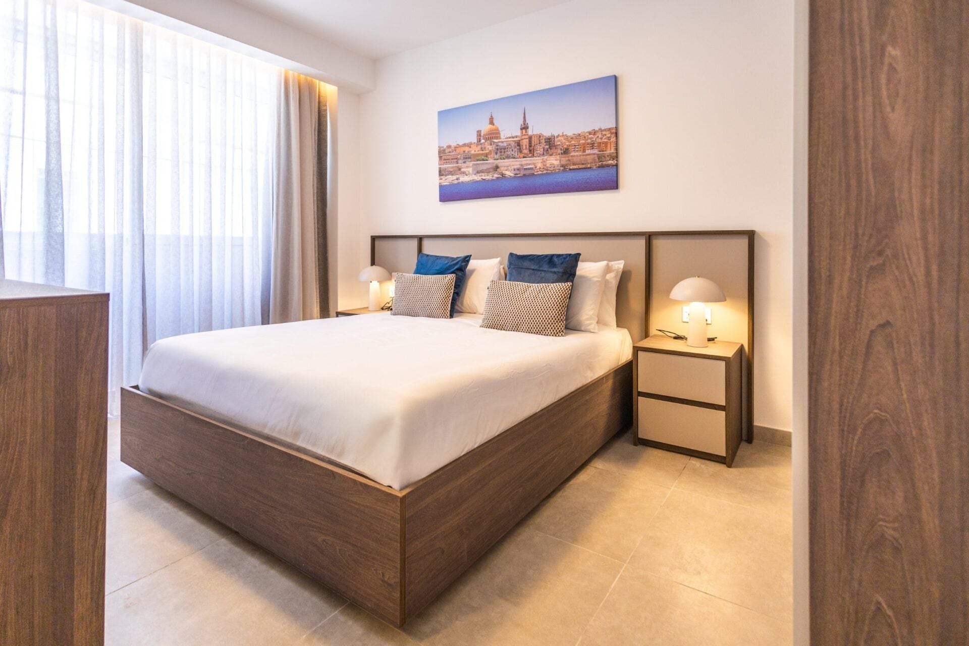 Un cuarto de hotel con una cama grande, dos mesitas de noche con lámparas, un cuadro de paisaje urbano en la pared y vistas de la ciudad a través de la ventana.