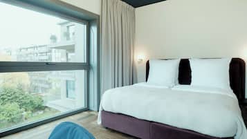 Habitación de hotel con un ventanal grande, una cama con ropa blanca y vistas a un edificio y árboles.