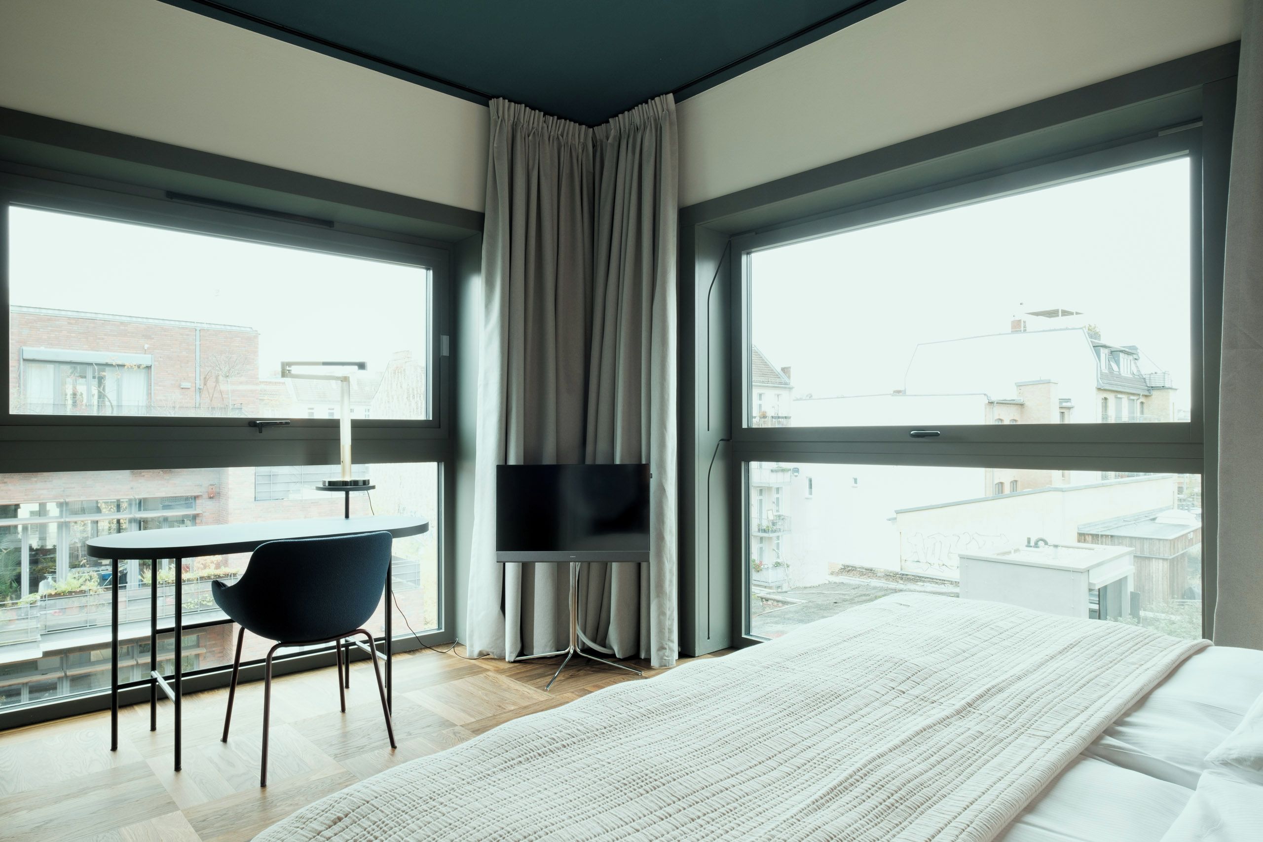 Una habitación de hotel moderna con una cama grande, un escritorio con silla, televisor de pantalla plana y ventanas amplias con vistas a la ciudad.