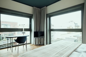 Ein modernes Hotelzimmer mit einem großen Bett, einem Schreibtisch mit Stuhl, einem Flachbildfernseher und großen Fenstern mit Blick auf die Stadt.