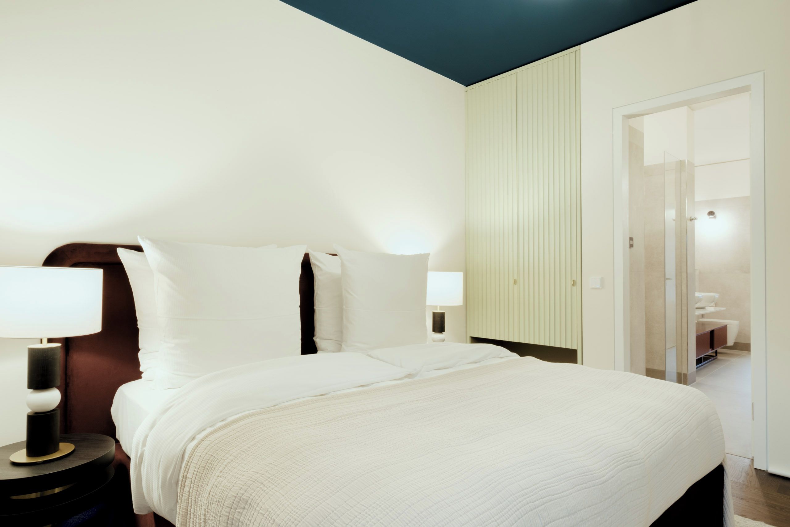 Habitación de hotel con una cama, mesitas de noche, una lámpara, una puerta que da a un baño y un techo azul.