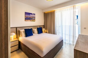 A hotel room with a large bed, a nightstand with a lamp, a painting on the wall, and a view of the outdoors through sheer curtains. - Spacious 1 Bedroom With Balcony (Msida)