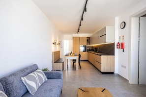 A modern living room with a sofa, a coffee table, and a dining area with a table and chairs. A kitchen with wooden cabinets and a built-in oven is visible in the background. - Relaxing 1BR Escape in Msida (Msida)