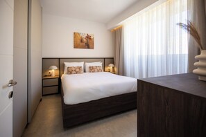 Ein Hotelzimmer mit einem großen Bett, zwei Nachttischen mit Lampen, einem Gemälde an der Wand und Blick ins Freie durch transparente Vorhänge.