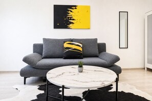 Een moderne woonkamer met een grijze bank, een ronde salontafel, een zwart-wit kleed, een abstract schilderij in geel en zwart, en een hoge spiegel.