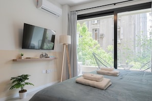 Une chambre d’hôtel moderne équipée d’un lit, d’un téléviseur à écran plat, d’une plante en pot, d’un vélo et d’un balcon offrant une vue sur les bâtiments et la verdure.