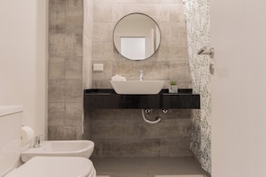 Une salle de bain avec un lavabo blanc, un miroir rond, des toilettes et un rideau de douche orné d’un motif de feuilles.
