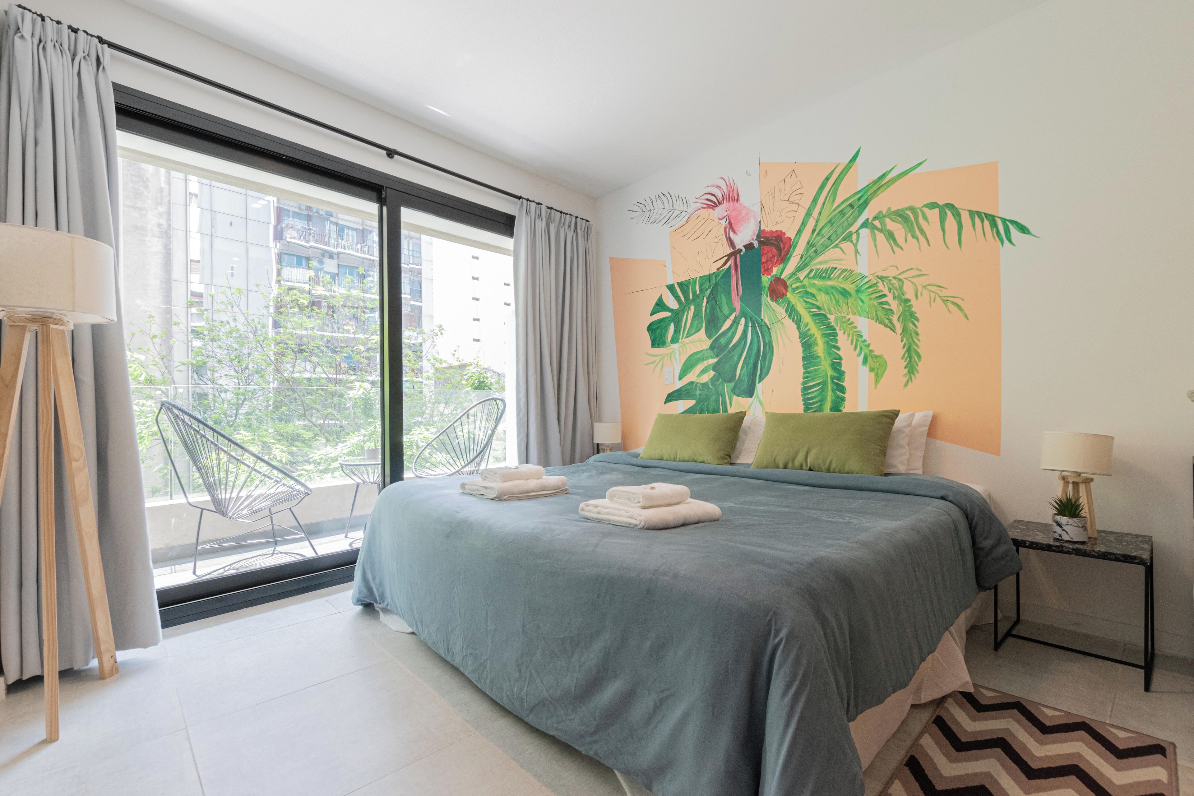 A modern hotel room with a large bed, a view of the city through floor-to-ceiling windows, a balcony with a chair, and a tropical-themed wall mural.