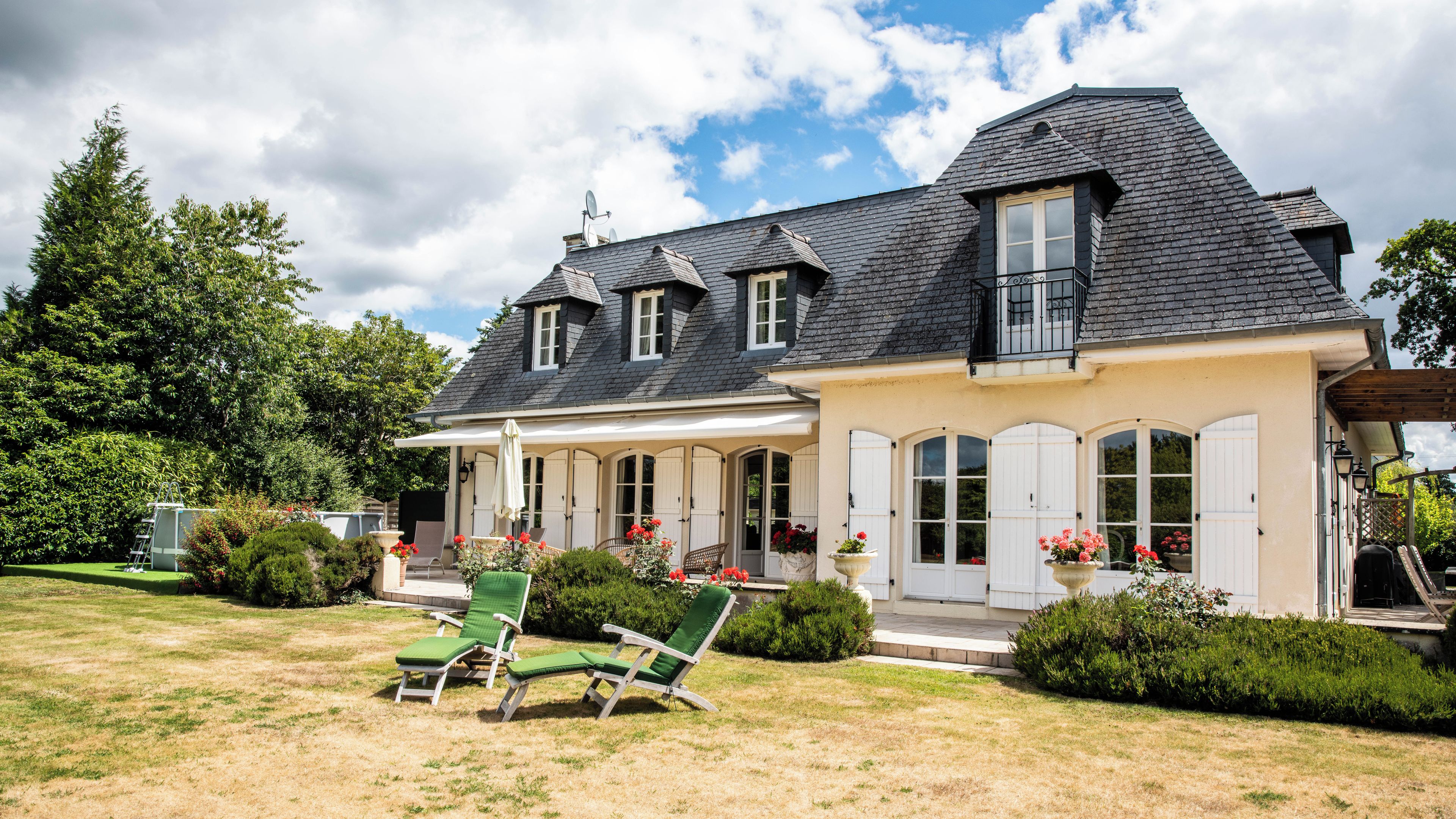 A two-story house with a covered porch, a lawn with green chairs, and a garden with flowers.
