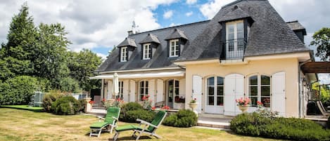 A two-story house with a covered porch, a lawn with green chairs, and a garden with flowers.