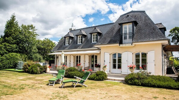 A two-story house with a covered porch, a lawn with green chairs, and a garden with flowers.