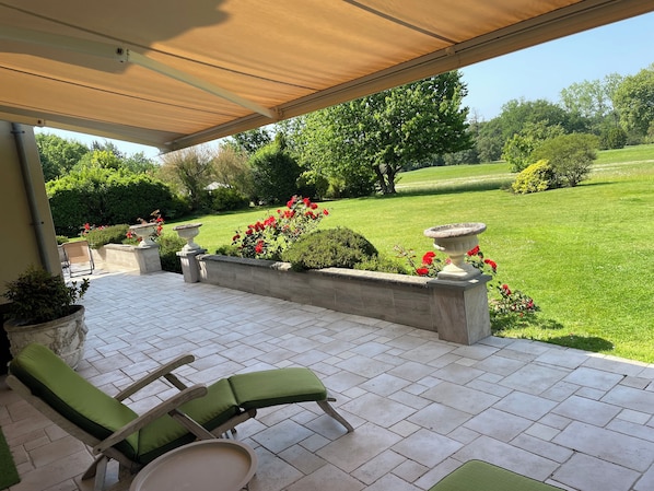 An outdoor patio with a covered seating area, a lawn, and a garden with flowers and shrubs.