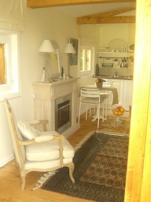 A room with a fireplace, a white chair, a dining table with chairs, and a kitchen area with a sink and cabinets.