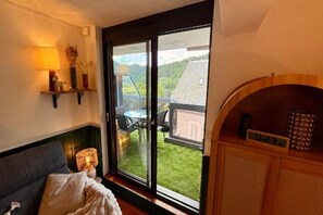 A room with a bed, a small table, a chair, a window with a view of a green landscape, and a wall-mounted shelf with decorative items. - Renovated apartment with balcony (Mont-Dore)