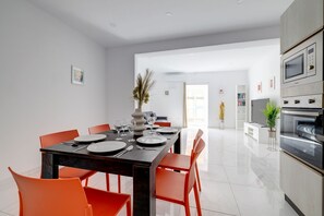 Cozinha moderna com forno embutido e micro-ondas, área de jantar com mesa de madeira escura e cadeiras laranja, e sala de estar ampla e iluminada com sofá e uma grande janela.