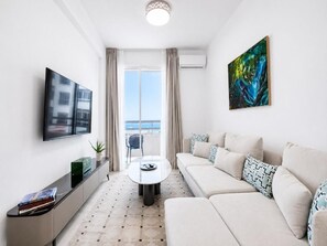 A modern living room with a sofa, a coffee table, a TV, and a painting. - Gardens Coastal Apt by TrulyCyprus (Lemesos)