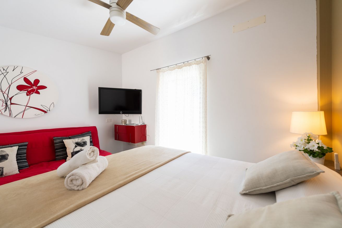 Habitación de hotel con cama, sofá rojo, televisor de pantalla plana, ventilador de techo y ventana con cortinas.