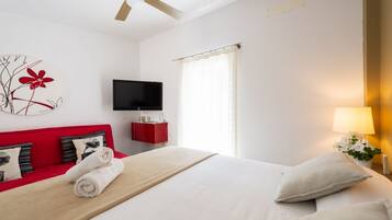 Habitación de hotel con cama, sofá rojo, televisor de pantalla plana, ventilador de techo y ventana con cortinas.