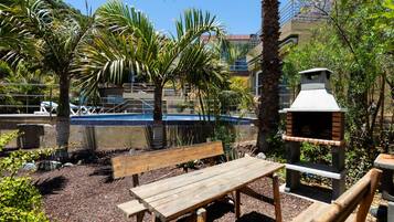 Un área de patio al aire libre con un banco de madera, una parrilla y una piscina de fondo.