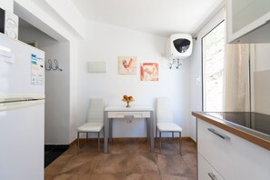 Una pequeña cocina con una mesa blanca, dos sillas, un frutero y una ventana con vistas.