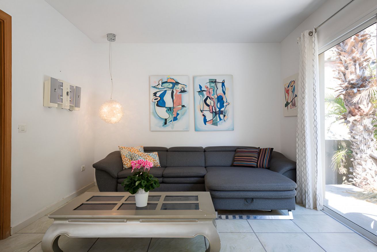 Een moderne woonkamer met een grijze bank, een glazen salontafel, abstracte kunst aan de muur en een plafondlamp.