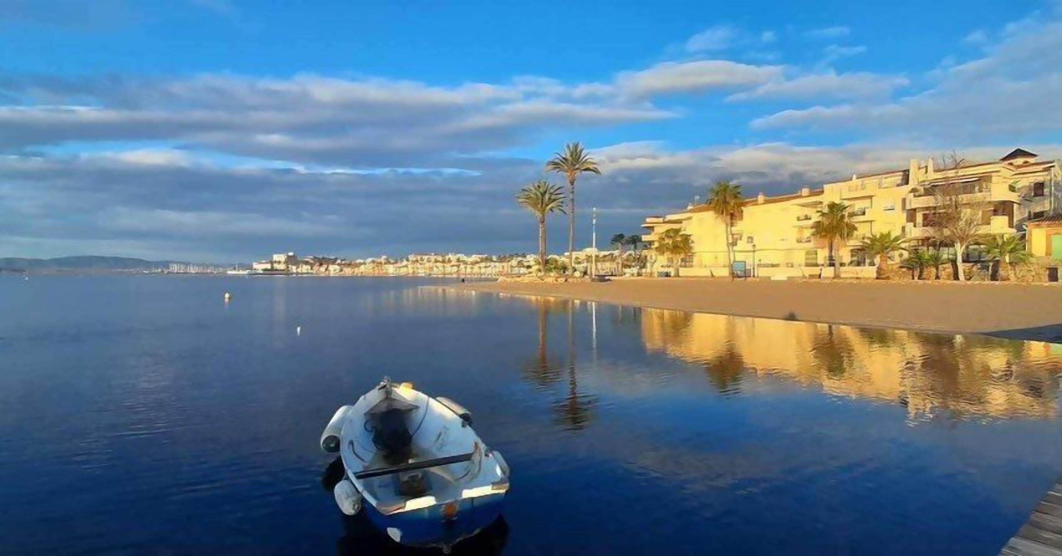 Un plan d’eau paisible, avec un bateau au premier plan, une plage et des bâtiments en arrière-plan.