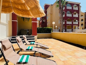 A sunlit outdoor seating area with lounge chairs and a thatched umbrella, adjacent to a building with balconies and a palm tree. - Casa Marc Angelo by Rental Hero (Alcazares, Los)