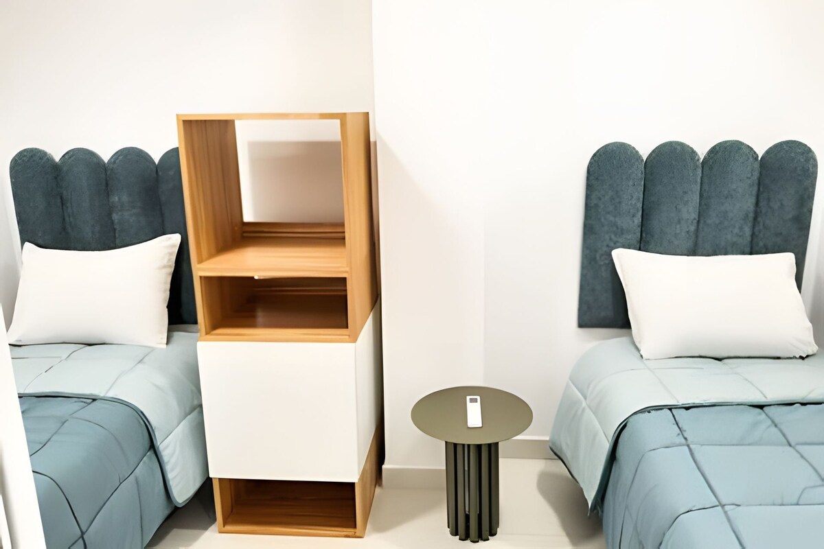 Una habitación con dos camas, cada una con cabecera, una mesita pequeña y una unidad estantería de madera empotrada.