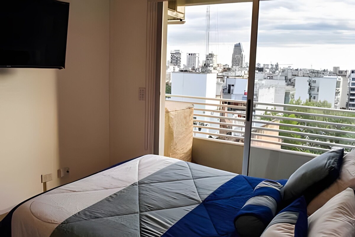 Un dormitorio con cama, ventana con vista a la ciudad y televisor montado en la pared.