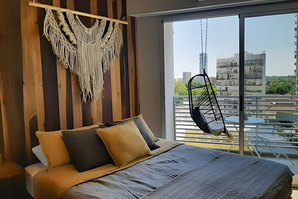 Un dormitorio con cama, almohadas, un colgador de macramé y vistas a edificios a través de una ventana.
