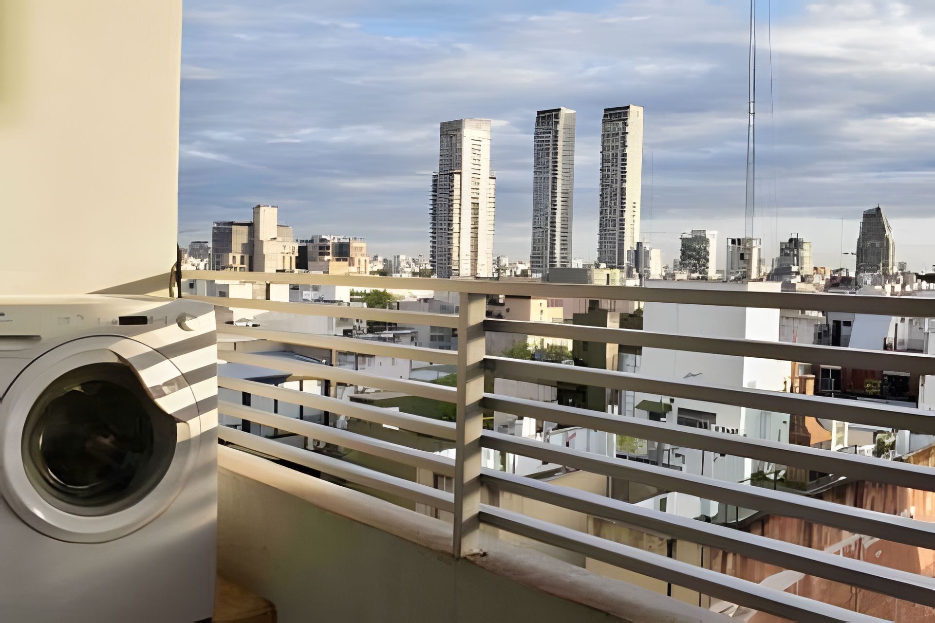 A balcony with a washing machine, overlooking a cityscape with multiple high-rise buildings.