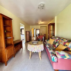 A living room with a sofa, a wooden cabinet, a round table with a floral tablecloth, and a ceiling light fixture. - Casa Nalon 2 by Rental Hero (Alcazares, Los)