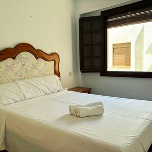 A neatly made bed with a white bedspread and a rolled towel on top, a wooden headboard with a floral pattern, a small wooden nightstand, and a window with blinds. - Casa Nalon 2 by Rental Hero (Alcazares, Los)