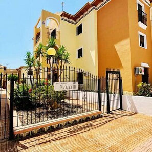 A gated entrance with a sign reading 'Playa III' and a paved pathway leading to a building with a yellow facade and a balcony. - Casa Playa by Rental Hero (Alcazares, Los)