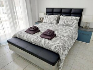 A neatly made bed with a leaf-patterned bedspread, two folded towels, a bench at the foot of the bed, and bedside tables with lamps. - Casa Palma by Rental Hero (Alcazares, Los)